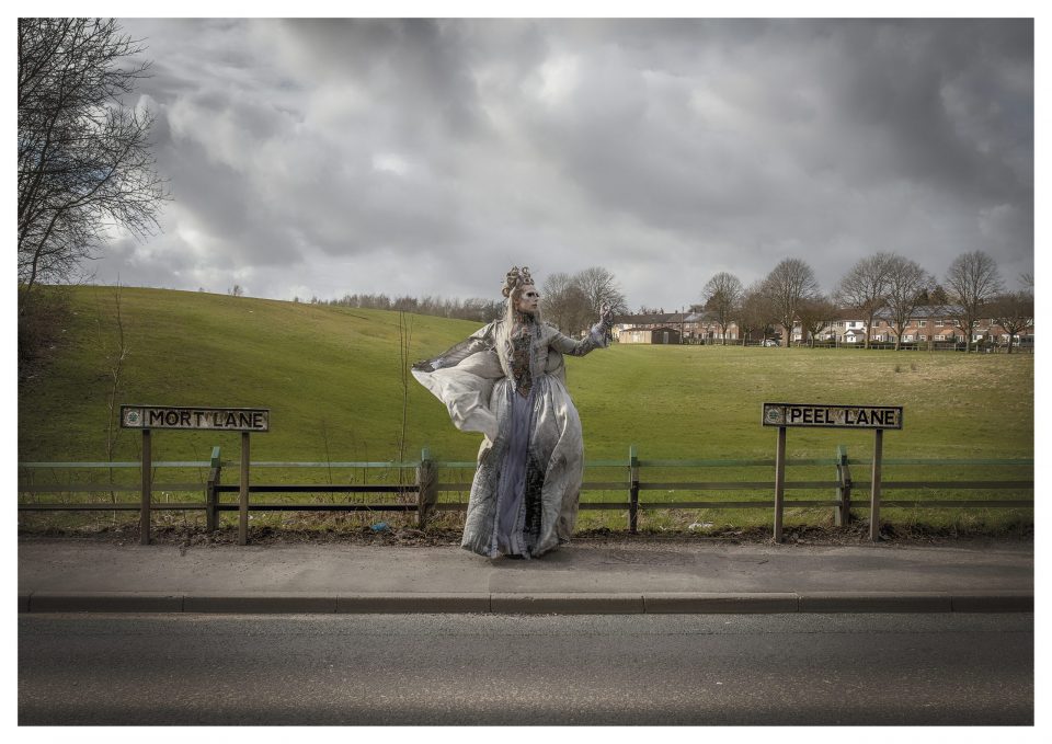 Cheddar Gorgeous stands on the pavement of peel lane with a field behind her, dressed in a grey gown and veil